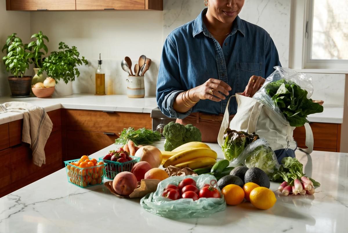 Woman unpacking fresh groceries, fruits, and leafy vegetables from a canvas tote bag onto a kitchen island. Woman unpacking fresh groceries, fruits, and leafy vegetables from a canvas tote bag onto a kitchen island.