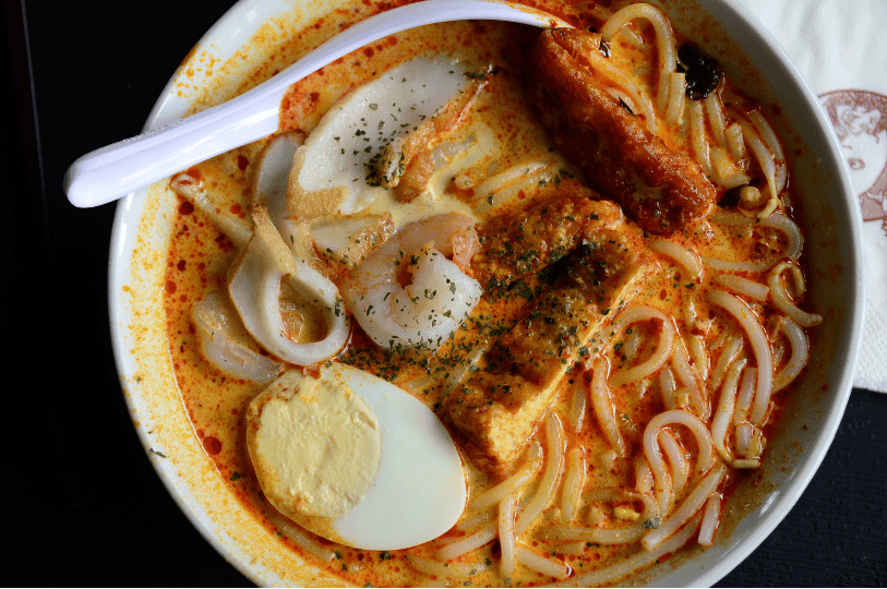 A top-down close-up of laksa in a white bowl, showcasing thick rice noodles in a vibrant coconut curry broth, topped with shrimp, sliced fish cake, fried tofu, and half a hard-boiled egg. A top-down close-up of laksa in a white bowl, showcasing thick rice noodles in a vibrant coconut curry broth, topped with shrimp, sliced fish cake, fried tofu, and half a hard-boiled egg.