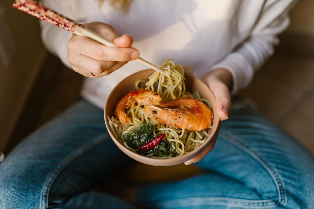 A person sits cross-legged holding a bowl of noodles topped with cooked shrimp, seaweed, and a red chili pepper. They use pattern-handled chopsticks to lift a portion of noodles from the dish. A person sits cross-legged holding a bowl of noodles topped with cooked shrimp, seaweed, and a red chili pepper. They use pattern-handled chopsticks to lift a portion of noodles from the dish.