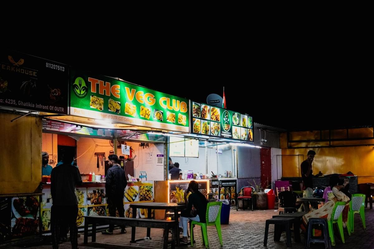 A brightly lit street food stall named "The Veg Club" operates at night, displaying colorful food photos on its green signage. Customers stand at the counter and sit at nearby outdoor tables while staff prepare food inside the stall. A brightly lit street food stall named "The Veg Club" operates at night, displaying colorful food photos on its green signage. Customers stand at the counter and sit at nearby outdoor tables while staff prepare food inside the stall.