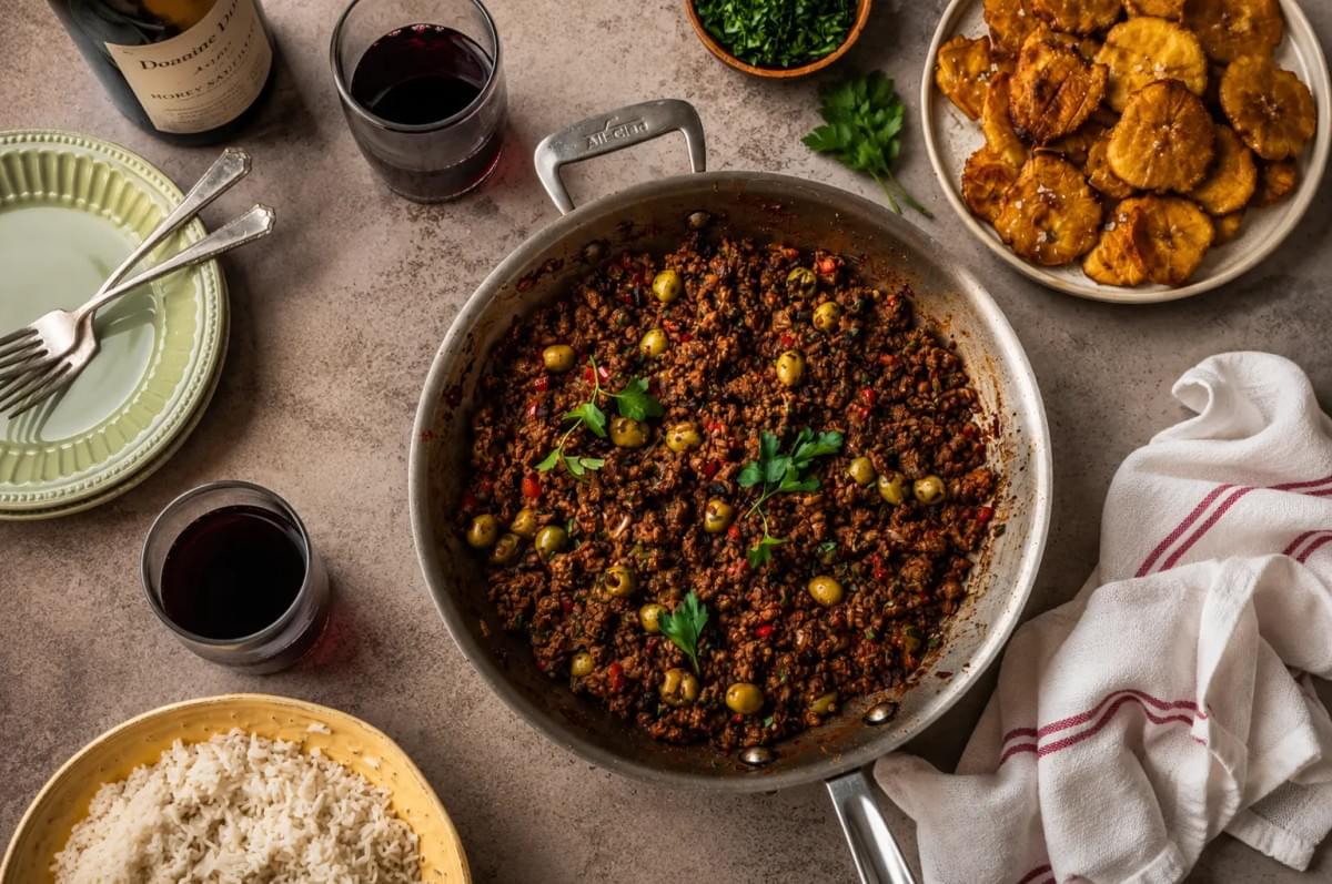 Top-down view of a silver skillet filled with seasoned picadillo and green olives, surrounded by plates of white rice, fried plantains, and wine glasses. Top-down view of a silver skillet filled with seasoned picadillo and green olives, surrounded by plates of white rice, fried plantains, and wine glasses.