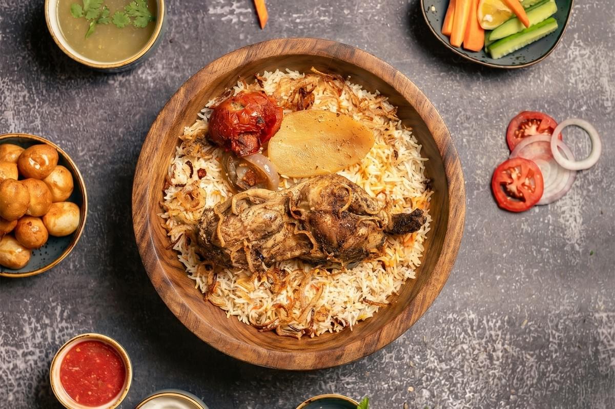 Overhead view of authentic Chicken Mandi rice served in a large wooden bowl, garnished with fried onions, a roasted tomato, and a potato slice, surrounded by side dishes and fresh vegetables Overhead view of authentic Chicken Mandi rice served in a large wooden bowl, garnished with fried onions, a roasted tomato, and a potato slice, surrounded by side dishes and fresh vegetables