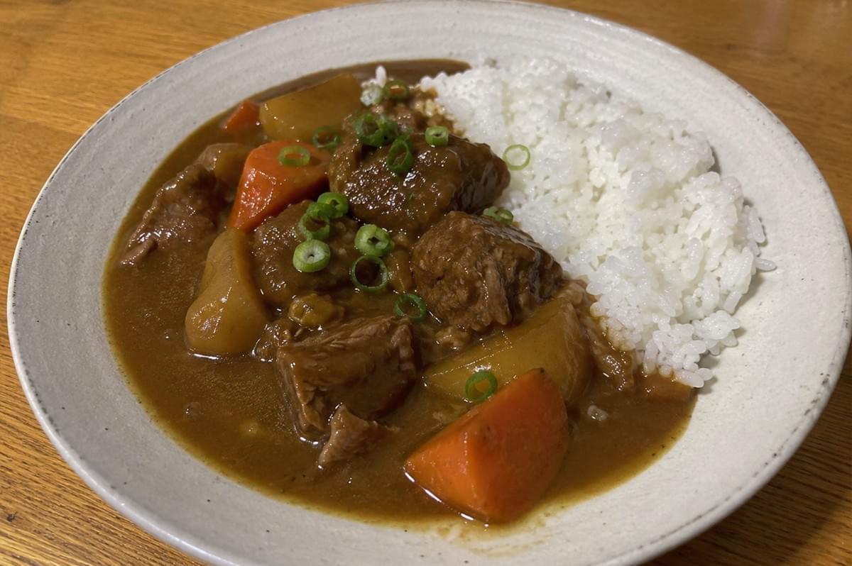 A top-down, slightly angled shot of a comforting bowl of Japanese beef curry served alongside a neat portion of fluffy white rice in a speckled, light gray ceramic bowl. The rich, thick, glossy brown curry sauce is studded with large, tender chunks of braised beef, vibrant orange carrot wedges, and soft, rounded pieces of potato. A scattering of freshly chopped green onions adds a bright pop of color over the center of the dish, all resting on a warm, natural wooden table. A top-down, slightly angled shot of a comforting bowl of Japanese beef curry served alongside a neat portion of fluffy white rice in a speckled, light gray ceramic bowl. The rich, thick, glossy brown curry sauce is studded with large, tender chunks of braised beef, vibrant orange carrot wedges, and soft, rounded pieces of potato. A scattering of freshly chopped green onions adds a bright pop of color over the center of the dish, all resting on a warm, natural wooden table.