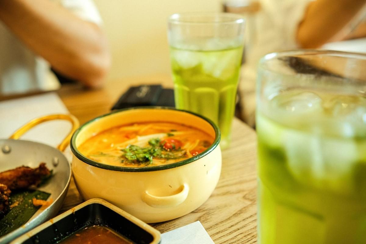 A bowl of savory soup topped with fresh herbs sits on a wooden dining table alongside tall glasses of iced green tea. Surrounding dishes and blurred diners complete the cozy restaurant setting. A bowl of savory soup topped with fresh herbs sits on a wooden dining table alongside tall glasses of iced green tea. Surrounding dishes and blurred diners complete the cozy restaurant setting.