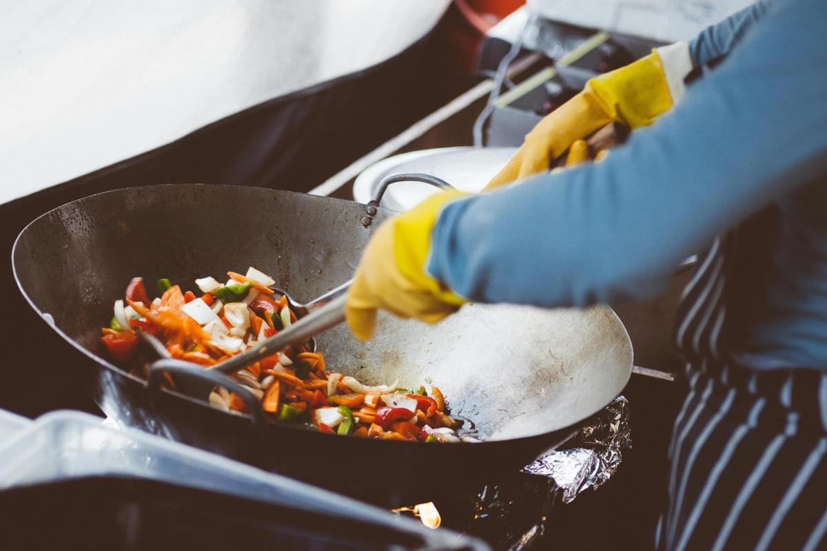 A cook wearing bright yellow gloves uses a large metal spoon to stir vibrant vegetables in a wok. The scene depicts a busy outdoor cooking environment with the wok heated over an open flame. A cook wearing bright yellow gloves uses a large metal spoon to stir vibrant vegetables in a wok. The scene depicts a busy outdoor cooking environment with the wok heated over an open flame.