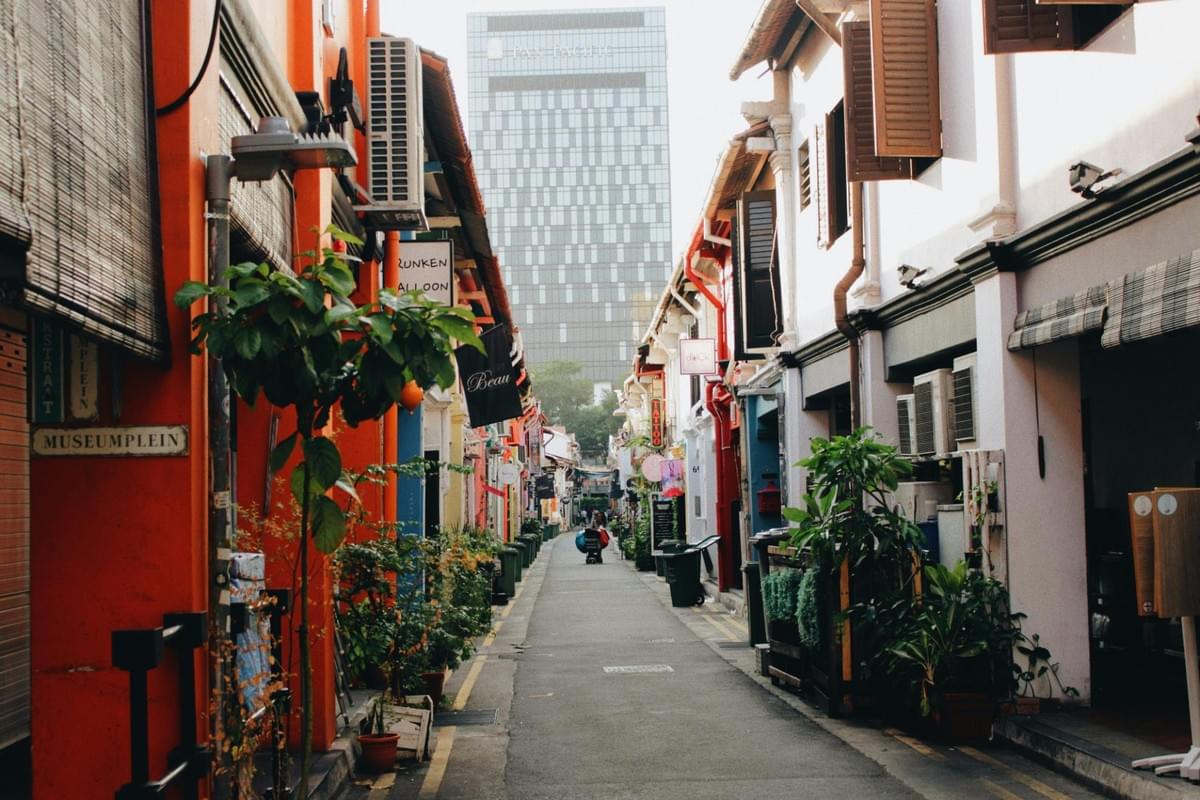 A narrow, charming alleyway is lined with colorful historic shophouses decorated with lush potted plants and wooden shutters. In the distance, a tall modern glass skyscraper looms over the traditional streetscape under a bright sky. A narrow, charming alleyway is lined with colorful historic shophouses decorated with lush potted plants and wooden shutters. In the distance, a tall modern glass skyscraper looms over the traditional streetscape under a bright sky.