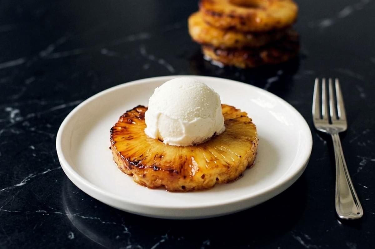 Caramelized pineapple ring topped with a scoop of vanilla ice cream on a white plate with fork, stacked pineapple rings in background Caramelized pineapple ring topped with a scoop of vanilla ice cream on a white plate with fork, stacked pineapple rings in background