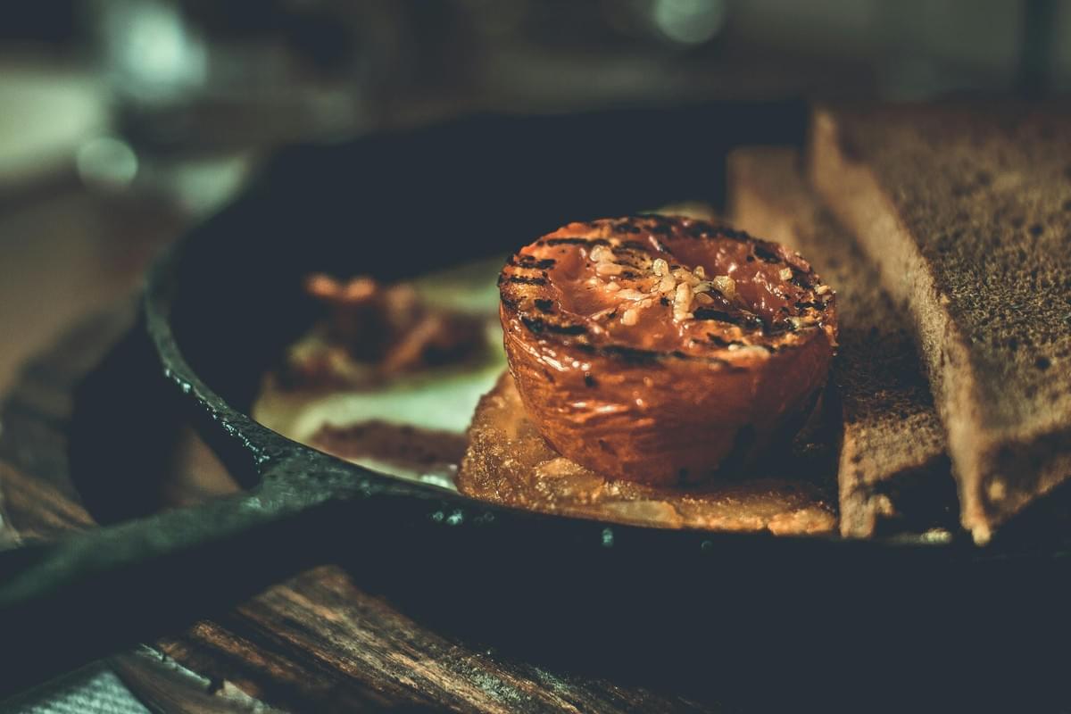 A close-up shot of a rustic breakfast served in a dark cast-iron skillet, featuring a grilled tomato half seasoned with herbs in the center. The tomato is flanked by thick slices of toasted brown bread, with a soft-focused background hint of eggs and bacon. A close-up shot of a rustic breakfast served in a dark cast-iron skillet, featuring a grilled tomato half seasoned with herbs in the center. The tomato is flanked by thick slices of toasted brown bread, with a soft-focused background hint of eggs and bacon.