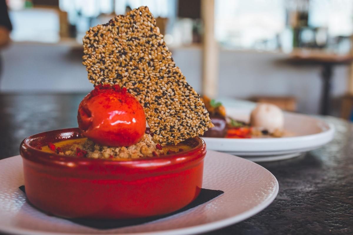 A close-up shot features a dessert served in a round, red ramekin, topped with a vibrant red scoop of sorbet and a tall, rectangular sesame seed crisp. In the blurred background, another dessert plate rests on a dark tabletop within a softly lit restaurant setting. A close-up shot features a dessert served in a round, red ramekin, topped with a vibrant red scoop of sorbet and a tall, rectangular sesame seed crisp. In the blurred background, another dessert plate rests on a dark tabletop within a softly lit restaurant setting.