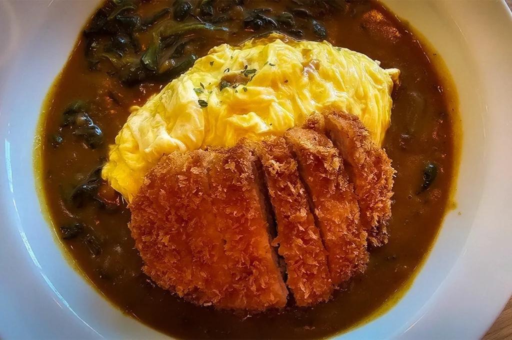 A white bowl filled with Japanese curry sauce featuring a mound of soft, fluffy swirled omelet and a crispy, sliced golden-brown katsu cutlet. Dark green spinach is mixed into the rich curry sauce surrounding the omelet and meat. A white bowl filled with Japanese curry sauce featuring a mound of soft, fluffy swirled omelet and a crispy, sliced golden-brown katsu cutlet. Dark green spinach is mixed into the rich curry sauce surrounding the omelet and meat.