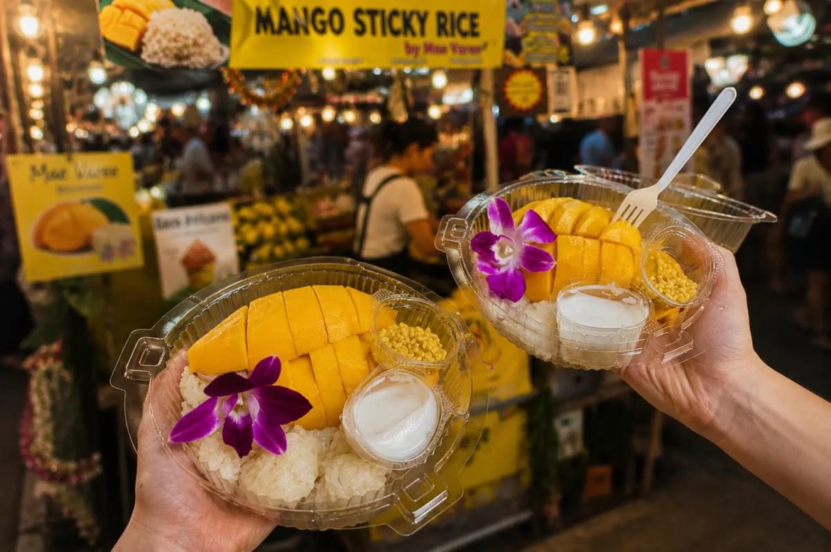Two plastic takeout containers of mango sticky rice held up in front of a night market stall. Two plastic takeout containers of mango sticky rice held up in front of a night market stall.