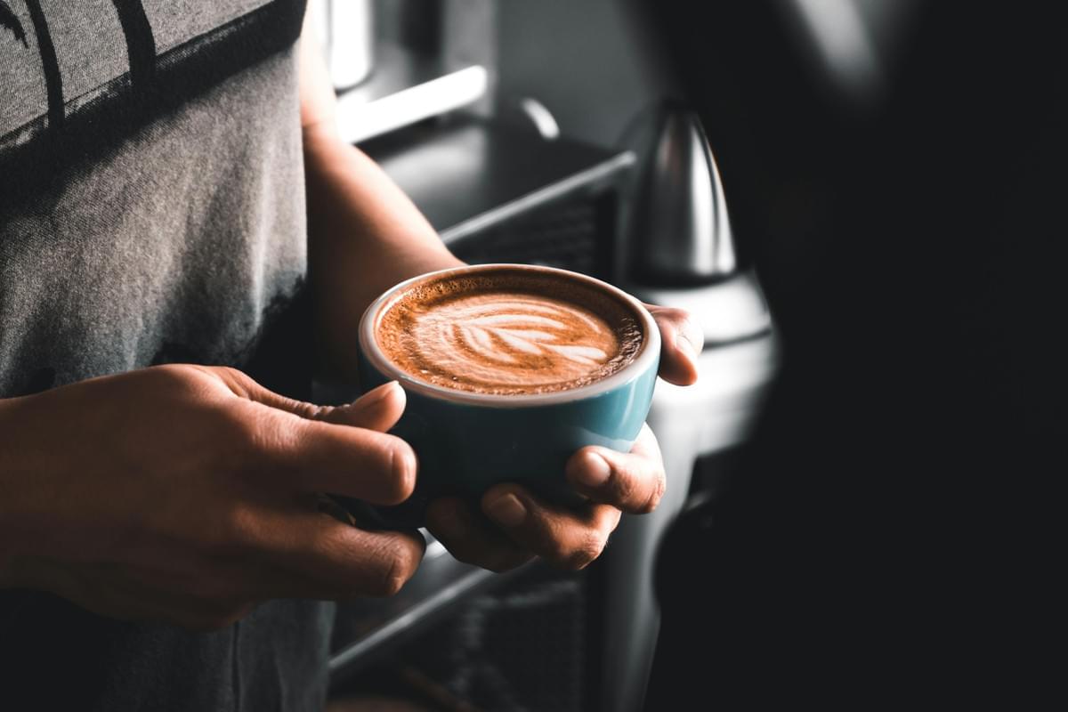 A close-up shot shows a person holding a blue ceramic mug filled with a fresh latte featuring detailed leaf latte art. The warm, moody lighting focuses on the hands cradling the cup against a dark, blurred background. A close-up shot shows a person holding a blue ceramic mug filled with a fresh latte featuring detailed leaf latte art. The warm, moody lighting focuses on the hands cradling the cup against a dark, blurred background.