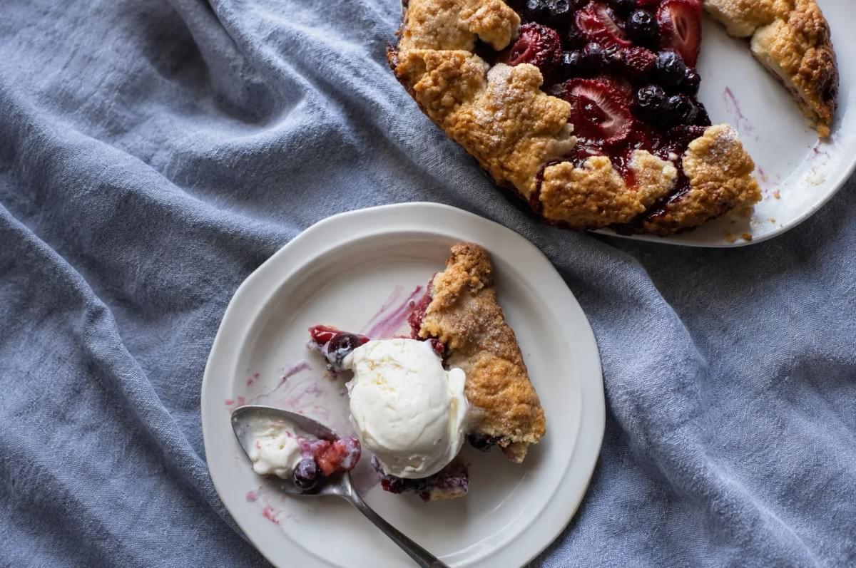 A slice of mixed berry galette topped with a scoop of vanilla ice cream on a plate next to the main tart. A slice of mixed berry galette topped with a scoop of vanilla ice cream on a plate next to the main tart.