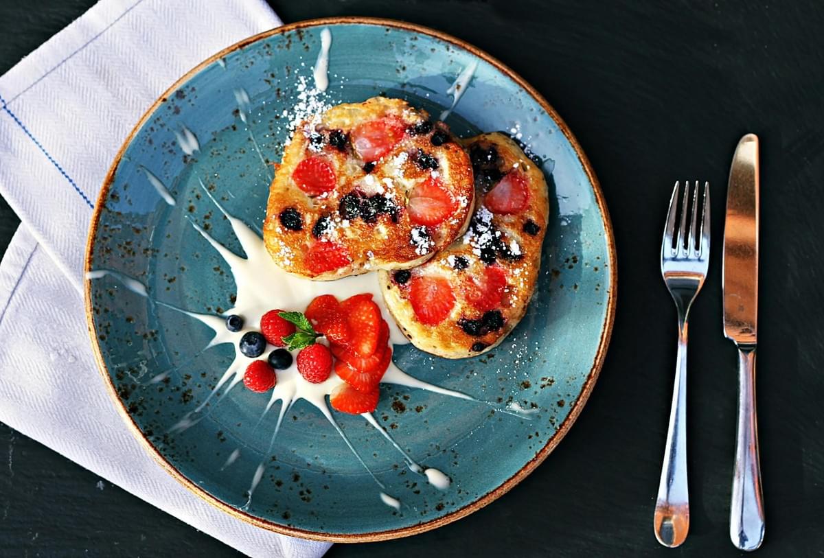 In the image, two golden-brown French toast slices topped with fresh strawberries, blueberries, and powdered sugar are served on a speckled blue plate. The plate is artistically decorated with a yogurt drizzle and a side of fresh berries, positioned next to a silver fork and knife on a dark surface. In the image, two golden-brown French toast slices topped with fresh strawberries, blueberries, and powdered sugar are served on a speckled blue plate. The plate is artistically decorated with a yogurt drizzle and a side of fresh berries, positioned next to a silver fork and knife on a dark surface.