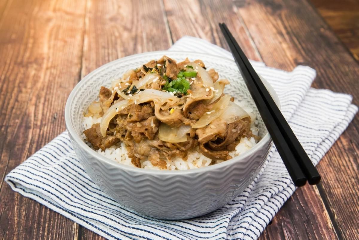 Gyudon beef rice bowl sprinkled with sesame seeds and green onions on a blue-lined napkin. Gyudon beef rice bowl sprinkled with sesame seeds and green onions on a blue-lined napkin.