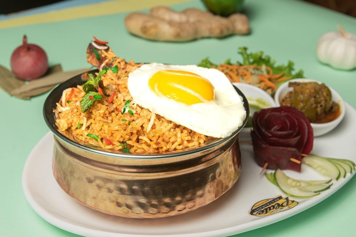 A metal bowl filled with seasoned orange rice and a chicken drumstick is topped with a sunny-side-up egg on a restaurant serving platter. The dish is accompanied by cucumber slices, a decorative beetroot rose, and small side bowls, set against a light green background with garlic, ginger, and onion. A metal bowl filled with seasoned orange rice and a chicken drumstick is topped with a sunny-side-up egg on a restaurant serving platter. The dish is accompanied by cucumber slices, a decorative beetroot rose, and small side bowls, set against a light green background with garlic, ginger, and onion.
