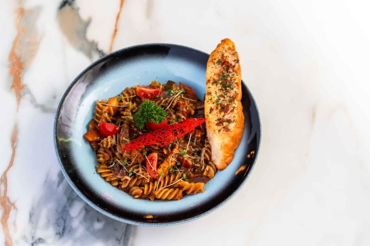 A top-down view shows a bowl of fusilli pasta tossed in a rich, dark sauce and garnished with cherry tomatoes, fresh herbs, and a red decorative tuile. A long piece of seasoned garlic bread rests on the side of the blue and black ceramic bowl, which sits on a white marble surface. A top-down view shows a bowl of fusilli pasta tossed in a rich, dark sauce and garnished with cherry tomatoes, fresh herbs, and a red decorative tuile. A long piece of seasoned garlic bread rests on the side of the blue and black ceramic bowl, which sits on a white marble surface.