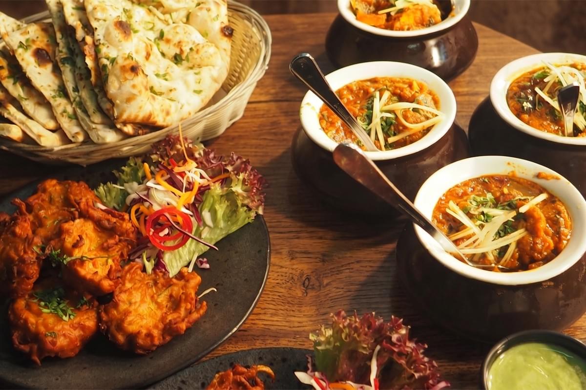 A spread of Pakistani cuisine featuring vegetable pakoras, garlic naan bread, and various curries in clay pots garnished with ginger and herbs. A spread of Pakistani cuisine featuring vegetable pakoras, garlic naan bread, and various curries in clay pots garnished with ginger and herbs.