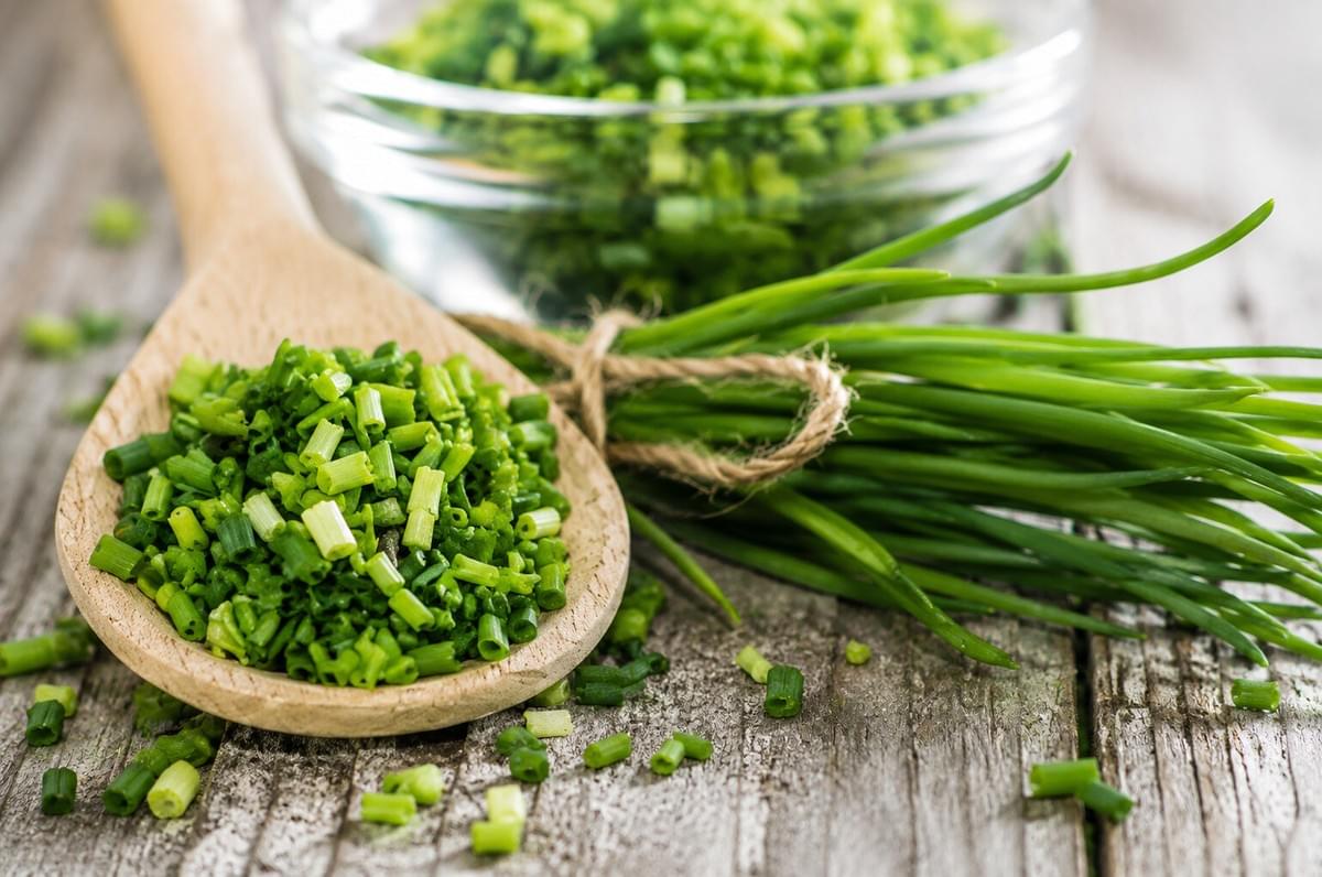 A wooden spoon filled with chopped chives next to a tied bundle of fresh chive stalks on a rustic wooden table. A wooden spoon filled with chopped chives next to a tied bundle of fresh chive stalks on a rustic wooden table.