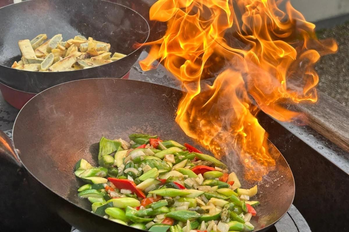 A vibrant stir-fry of green and red vegetables cooks in a large wok as dramatic, bright orange flames erupt from the side. In the background, a second wok holds lightly browned, cut pieces of food being prepared simultaneously. A vibrant stir-fry of green and red vegetables cooks in a large wok as dramatic, bright orange flames erupt from the side. In the background, a second wok holds lightly browned, cut pieces of food being prepared simultaneously.