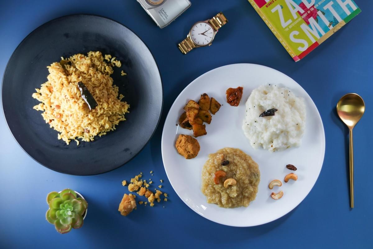 A top-down view shows a South Indian meal arranged on a vibrant blue background next to a watch, a book, and a small plant. A black plate holds spiced yellow rice, while a white plate features portions of curd rice, sweet pongal, roasted potatoes, and small accompaniments. A top-down view shows a South Indian meal arranged on a vibrant blue background next to a watch, a book, and a small plant. A black plate holds spiced yellow rice, while a white plate features portions of curd rice, sweet pongal, roasted potatoes, and small accompaniments.