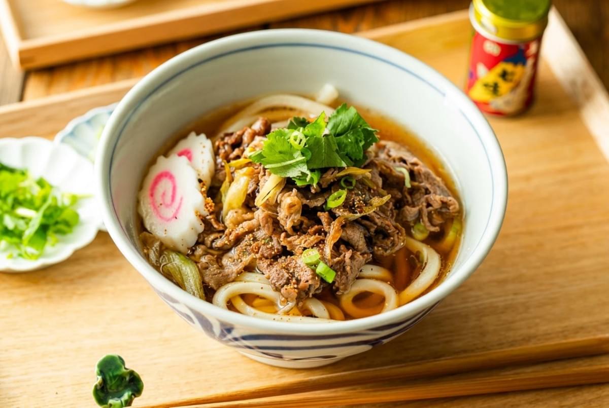 Bowl of beef udon noodle soup topped with fresh herbs and sliced fish cake on a wooden tray. Bowl of beef udon noodle soup topped with fresh herbs and sliced fish cake on a wooden tray.
