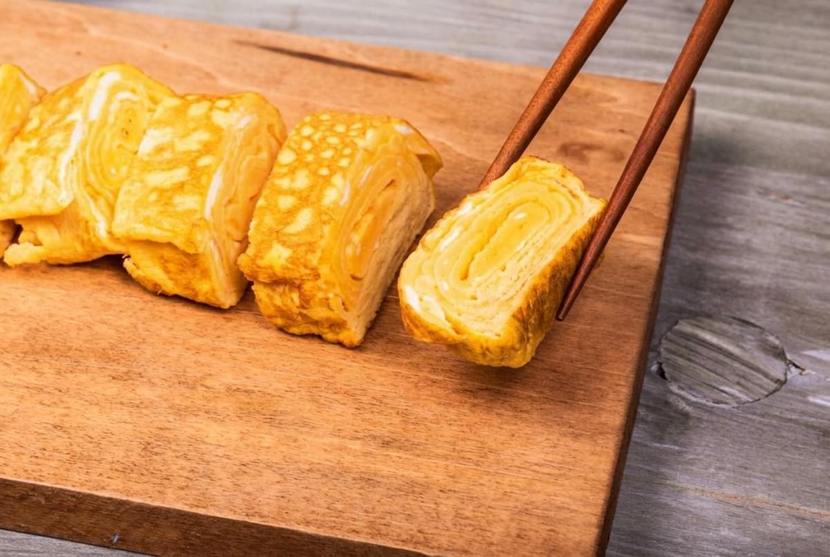 Chopsticks picking up a slice of rolled tamagoyaki omelet from a wooden serving board. Chopsticks picking up a slice of rolled tamagoyaki omelet from a wooden serving board.