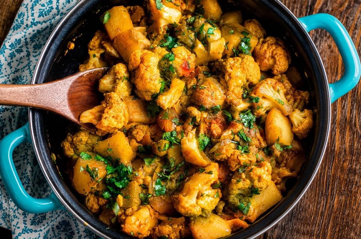 A wooden spoon stirring freshly cooked Aloo Gobi topped with chopped cilantro in a teal-handled pot. A wooden spoon stirring freshly cooked Aloo Gobi topped with chopped cilantro in a teal-handled pot.