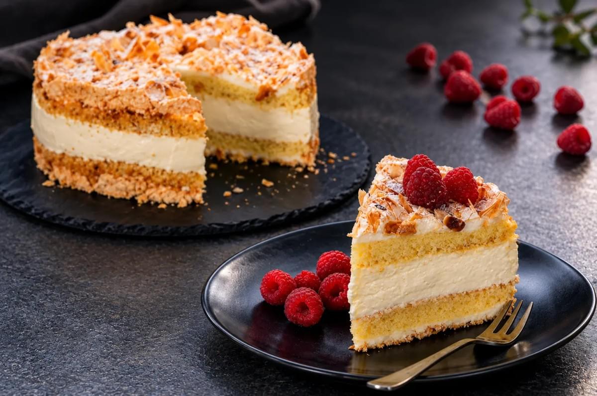 Slice of almond meringue cake with fresh raspberries on a black plate, next to the main cake. Slice of almond meringue cake with fresh raspberries on a black plate, next to the main cake.
