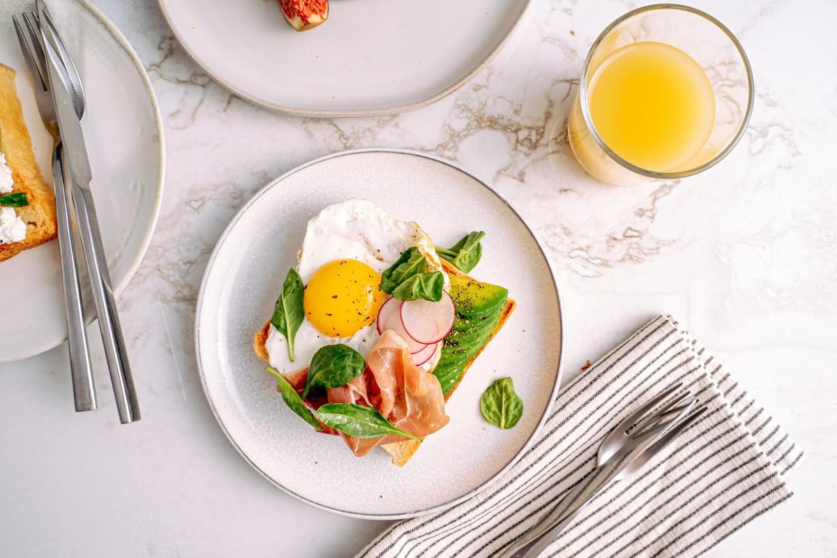 A top-down view of a bright breakfast scene featuring an open-faced sandwich topped with a sunny-side-up egg, sliced avocado, prosciutto, radishes, and spinach leaves on a white ceramic plate. The dish is set on a white marble tabletop alongside a glass of orange juice, utensils, and partial views of other breakfast plates. A top-down view of a bright breakfast scene featuring an open-faced sandwich topped with a sunny-side-up egg, sliced avocado, prosciutto, radishes, and spinach leaves on a white ceramic plate. The dish is set on a white marble tabletop alongside a glass of orange juice, utensils, and partial views of other breakfast plates.