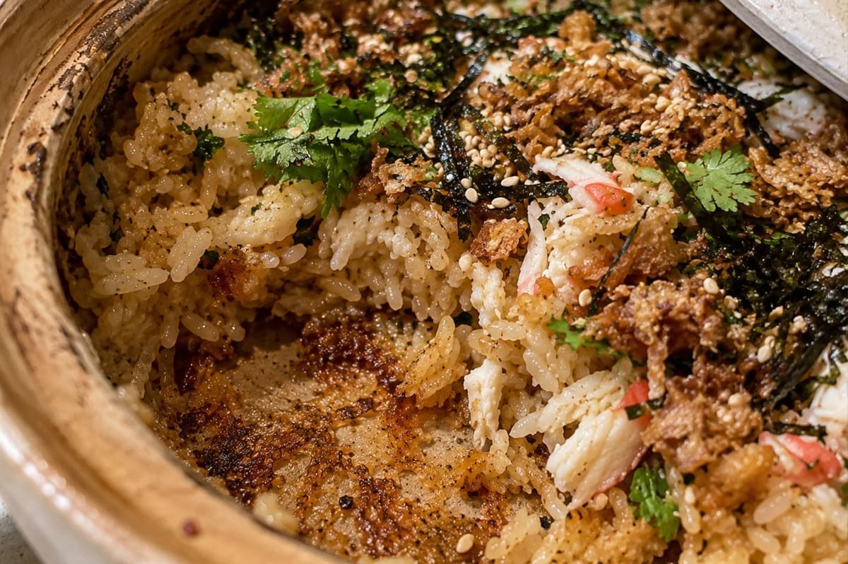 A close-up shot inside the earthenware claypot reveals a section where the rice has been scooped away to highlight the golden, caramelized crispy rice layer (nurungji or okoroge) formed along the bottom. Flaky white crab meat, toasted sesame seeds, shredded nori, crispy fried shallots, and fresh cilantro garnish the remaining seasoned rice inside the pot. A close-up shot inside the earthenware claypot reveals a section where the rice has been scooped away to highlight the golden, caramelized crispy rice layer (nurungji or okoroge) formed along the bottom. Flaky white crab meat, toasted sesame seeds, shredded nori, crispy fried shallots, and fresh cilantro garnish the remaining seasoned rice inside the pot.