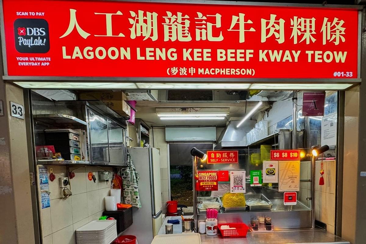Exterior of a small food stall with a bright red sign reading "Lagoon Leng Kee Beef Kway Teow." The stall is neat, offering self-service options. Exterior of a small food stall with a bright red sign reading "Lagoon Leng Kee Beef Kway Teow." The stall is neat, offering self-service options.