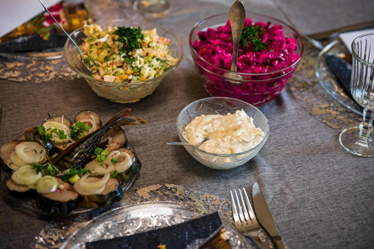 Several glass bowls and plates display an assortment of dishes on a grey tablecloth, including a vibrant purple beet salad and a creamy vegetable mix. In the foreground, slices of fish garnished with onion rings and green herbs are placed next to a small bowl of white dip and silver cutlery. Several glass bowls and plates display an assortment of dishes on a grey tablecloth, including a vibrant purple beet salad and a creamy vegetable mix. In the foreground, slices of fish garnished with onion rings and green herbs are placed next to a small bowl of white dip and silver cutlery.