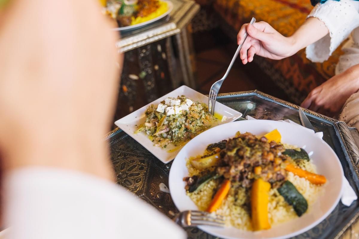 A close-up shot shows a hand using a fork to taste a fresh green appetizer topped with feta cheese. Next to it sits a large plate of couscous topped with cooked vegetables and seasoned meat on an ornate tray table. A close-up shot shows a hand using a fork to taste a fresh green appetizer topped with feta cheese. Next to it sits a large plate of couscous topped with cooked vegetables and seasoned meat on an ornate tray table.