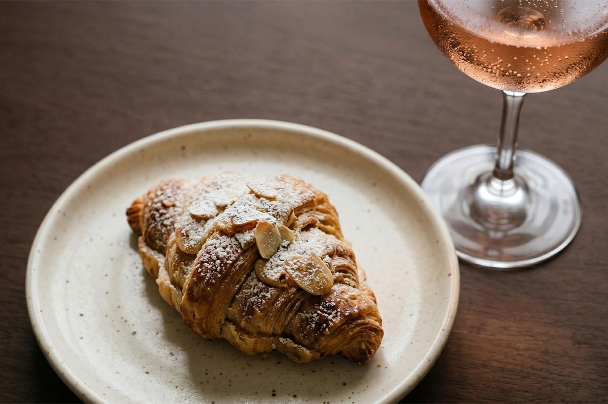 An almond croissant topped with powdered sugar on a plate next to a glass of sparkling rose. An almond croissant topped with powdered sugar on a plate next to a glass of sparkling rose.
