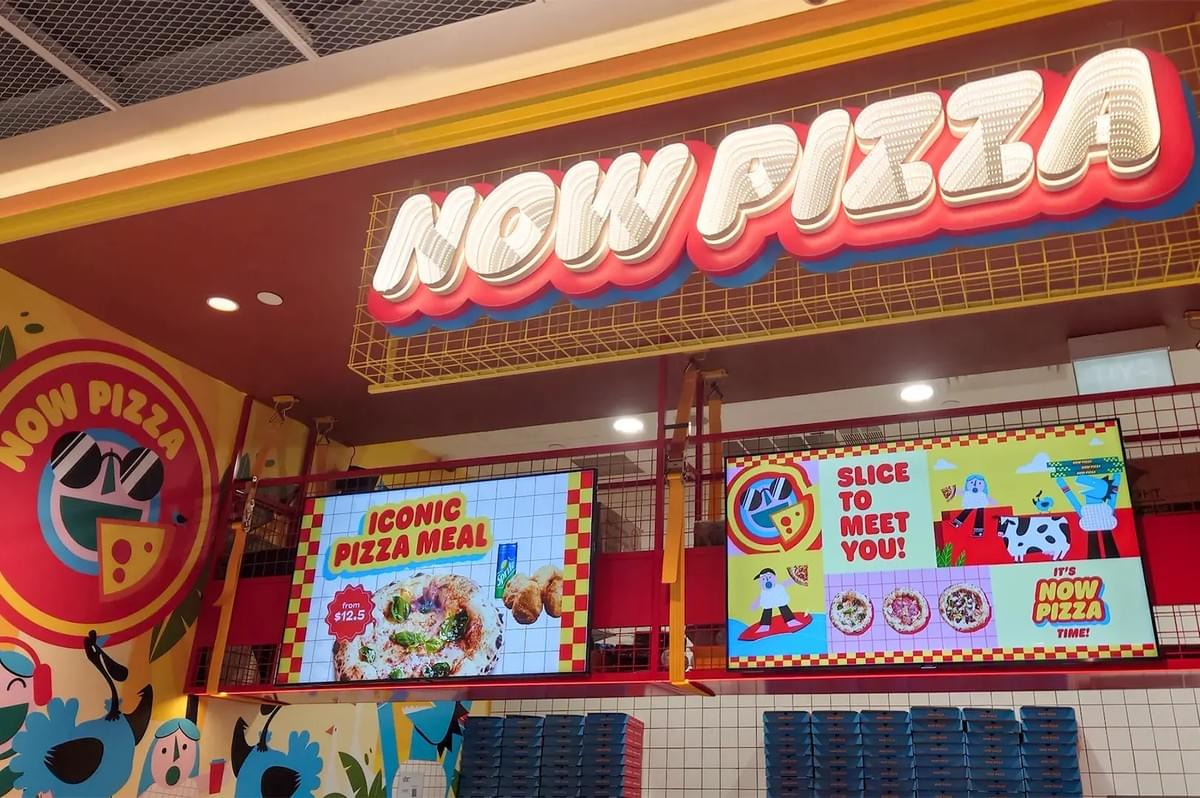 A colorful restaurant counter at Now Pizza featuring a vibrant neon sign and digital menu boards displaying an "Iconic Pizza Meal" and cartoon illustrations. A colorful restaurant counter at Now Pizza featuring a vibrant neon sign and digital menu boards displaying an "Iconic Pizza Meal" and cartoon illustrations.