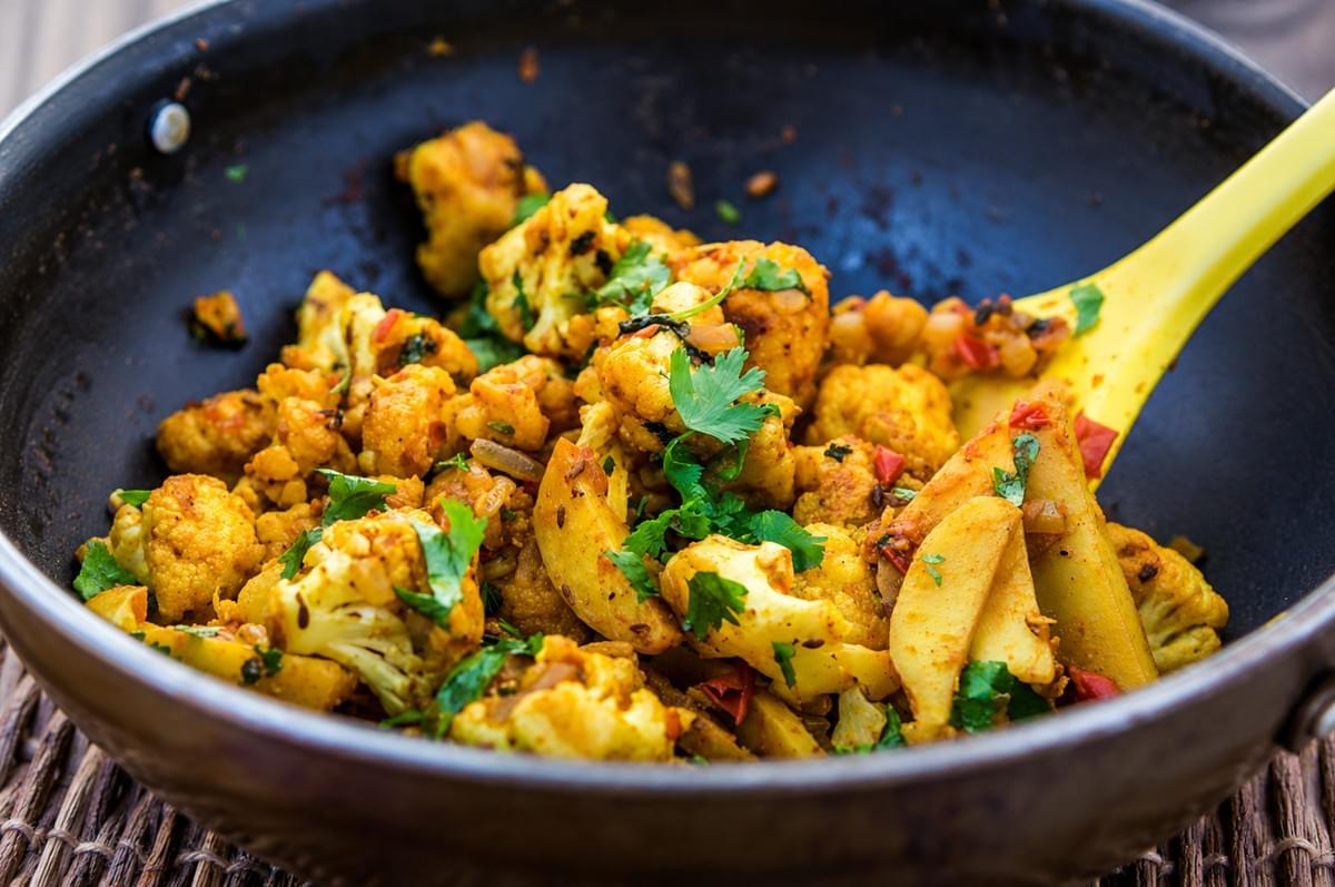 A yellow utensil stirring spiced cauliflower florets and potatoes with fresh cilantro in a dark wok. A yellow utensil stirring spiced cauliflower florets and potatoes with fresh cilantro in a dark wok.