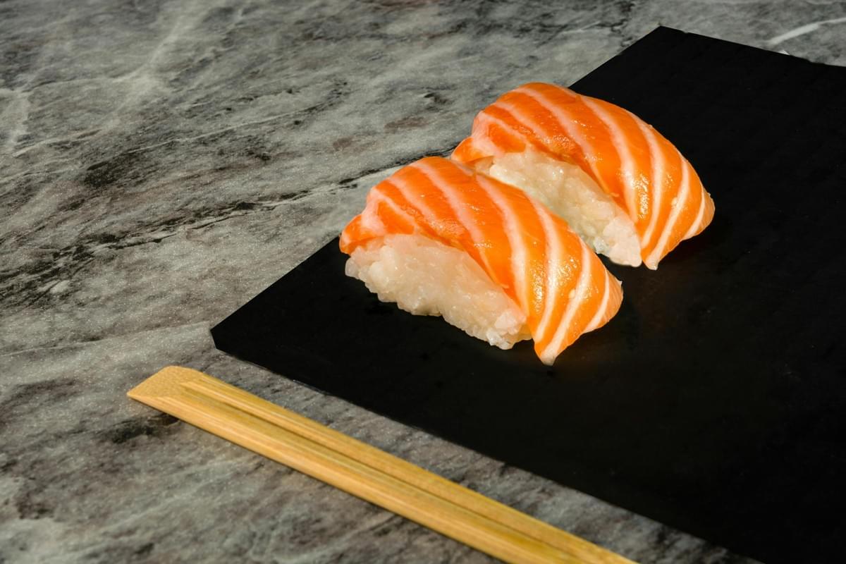 Two pieces of salmon nigiri with vibrant white marbling are served on a sleek black slate board. A pair of light wood chopsticks rests alongside the plate on a textured, grey marble-style surface. Two pieces of salmon nigiri with vibrant white marbling are served on a sleek black slate board. A pair of light wood chopsticks rests alongside the plate on a textured, grey marble-style surface.