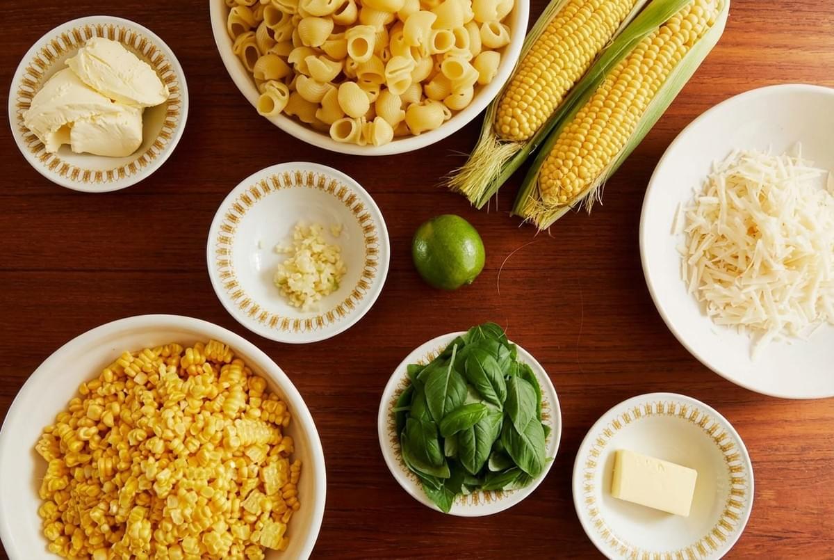 Top-down view of ingredients for sweet corn pasta, including raw cobs, kernels, shell pasta, butter, garlic, basil, and cheese. Top-down view of ingredients for sweet corn pasta, including raw cobs, kernels, shell pasta, butter, garlic, basil, and cheese.