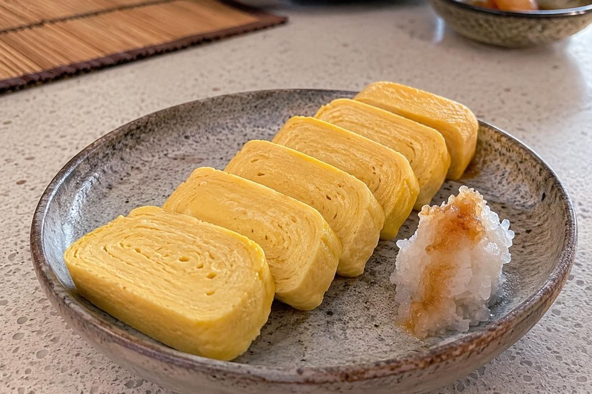 Five neat, golden slices of rolled Japanese omelet (tamagoyaki) are arranged diagonally on a textured grey ceramic plate alongside a small mound of grated daikon radish drizzled with soy sauce. Five neat, golden slices of rolled Japanese omelet (tamagoyaki) are arranged diagonally on a textured grey ceramic plate alongside a small mound of grated daikon radish drizzled with soy sauce.