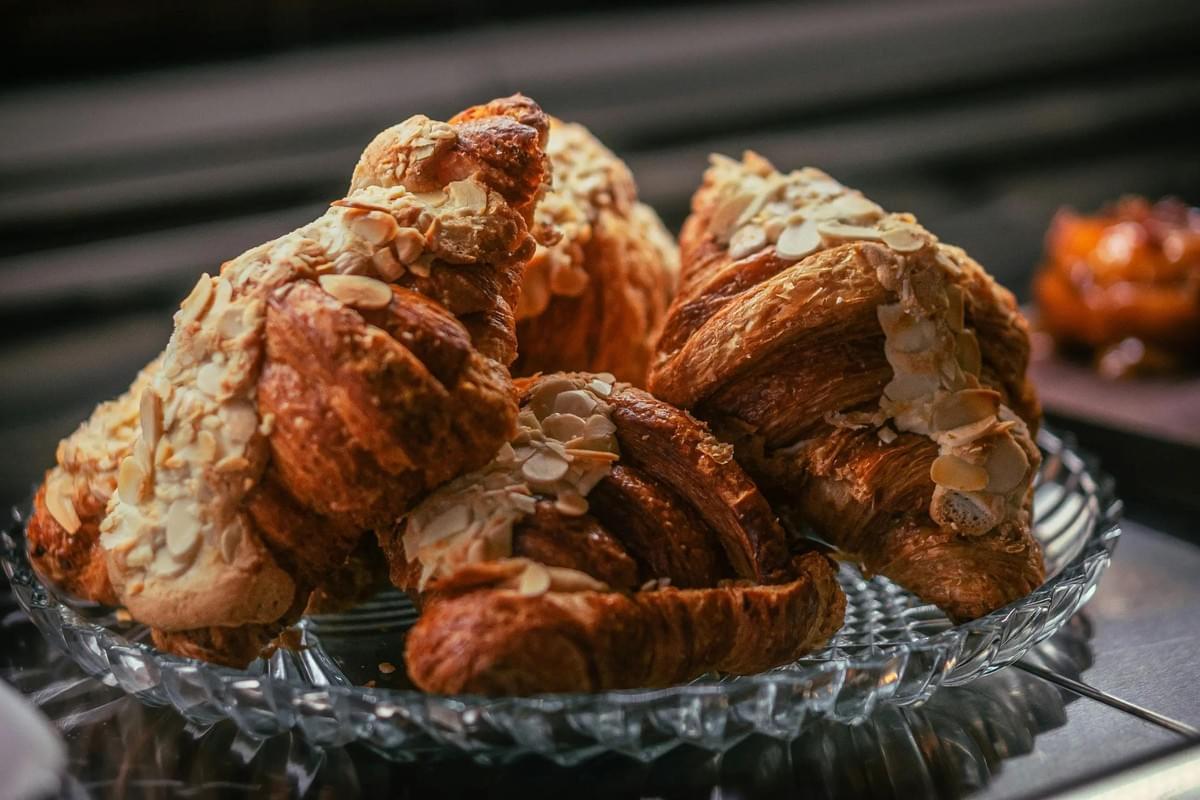 A close-up shot of several golden-brown, flaky almond croissants piled onto a decorative glass platter. In the file, the pastries are generously topped with sliced almonds and a light dusting of powdered sugar. A close-up shot of several golden-brown, flaky almond croissants piled onto a decorative glass platter. In the file, the pastries are generously topped with sliced almonds and a light dusting of powdered sugar.