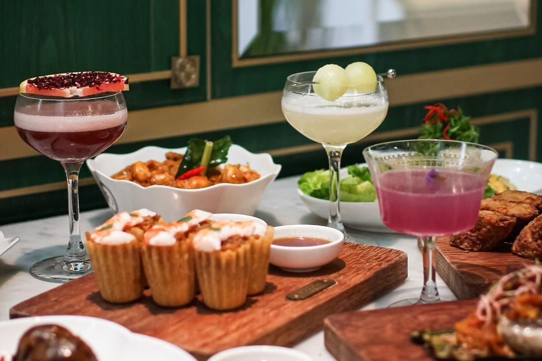 A table setting features an assortment of colorful craft cocktails and Southeast Asian-style appetizers, including kueh pie tee and curry dishes. The drinks are garnished with fresh fruit slice toppings, melon balls, and edible flowers. A table setting features an assortment of colorful craft cocktails and Southeast Asian-style appetizers, including kueh pie tee and curry dishes. The drinks are garnished with fresh fruit slice toppings, melon balls, and edible flowers.