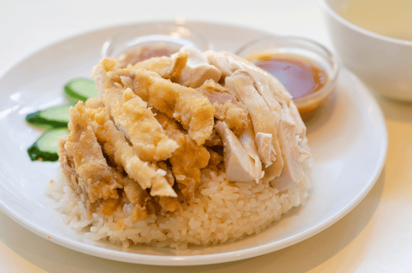 A plate of Khao Man Gai (Hainan Chicken Rice) featuring both poached and crispy fried chicken slices over seasoned rice, served with cucumber garnishes and dipping sauces. A plate of Khao Man Gai (Hainan Chicken Rice) featuring both poached and crispy fried chicken slices over seasoned rice, served with cucumber garnishes and dipping sauces.