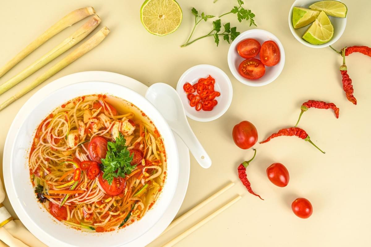 A top-down view of a bowl of spicy noodle soup garnished with fresh herbs, sliced tomatoes, and chilies. Surrounding the bowl on a light yellow background are fresh ingredients, including lemongrass stalks, lime wedges, cherry tomatoes, and red chili peppers. A top-down view of a bowl of spicy noodle soup garnished with fresh herbs, sliced tomatoes, and chilies. Surrounding the bowl on a light yellow background are fresh ingredients, including lemongrass stalks, lime wedges, cherry tomatoes, and red chili peppers.