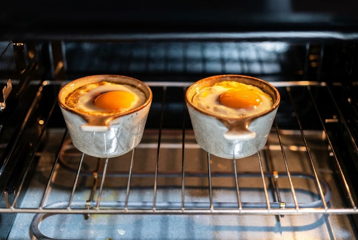 Two ceramic ramekins with raw eggs baking inside an oven on a wire rack. Two ceramic ramekins with raw eggs baking inside an oven on a wire rack.