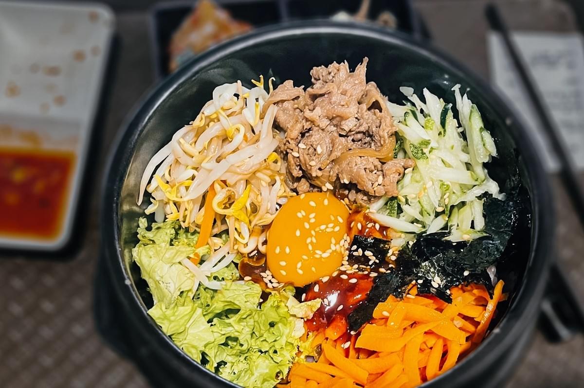 Top-down close-up shot of a Korean-style rice bowl with a raw egg yolk at the center, surrounded by sliced vegetables including bean sprouts, shredded carrots, lettuce, and cucumber, with marinated meat, seaweed, and sesame seeds in a black bowl on a dining table with blurred side dishes in the background. Top-down close-up shot of a Korean-style rice bowl with a raw egg yolk at the center, surrounded by sliced vegetables including bean sprouts, shredded carrots, lettuce, and cucumber, with marinated meat, seaweed, and sesame seeds in a black bowl on a dining table with blurred side dishes in the background.