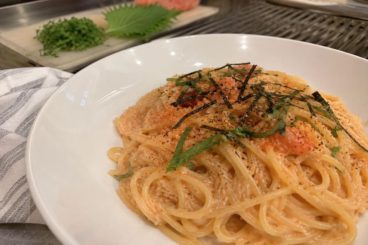 The image displays a close-up, high-angle view of a white bowl filled with creamy Mentaiko spaghetti, a popular Japanese-fusion pasta dish. The noodles are coated in a rich, pale orange sauce made from spicy cod roe, garnished with delicate strips of dark nori seaweed, finely sliced green shiso leaves, and a light dusting of red spice, likely shichimi togarashi. In the blurred background, a cutting board holds a fresh, green shiso leaf and a small pile of finely chopped herbs, while a small plastic container sits nearby, suggesting a clean and organized kitchen environment. A grey and white striped cloth is partially visible on the left side of the frame, providing a soft contrast to the smooth, bright white surface of the pasta bowl and the industrial-looking, ribbed metal countertop beneath it. The image displays a close-up, high-angle view of a white bowl filled with creamy Mentaiko spaghetti, a popular Japanese-fusion pasta dish. The noodles are coated in a rich, pale orange sauce made from spicy cod roe, garnished with delicate strips of dark nori seaweed, finely sliced green shiso leaves, and a light dusting of red spice, likely shichimi togarashi. In the blurred background, a cutting board holds a fresh, green shiso leaf and a small pile of finely chopped herbs, while a small plastic container sits nearby, suggesting a clean and organized kitchen environment. A grey and white striped cloth is partially visible on the left side of the frame, providing a soft contrast to the smooth, bright white surface of the pasta bowl and the industrial-looking, ribbed metal countertop beneath it.