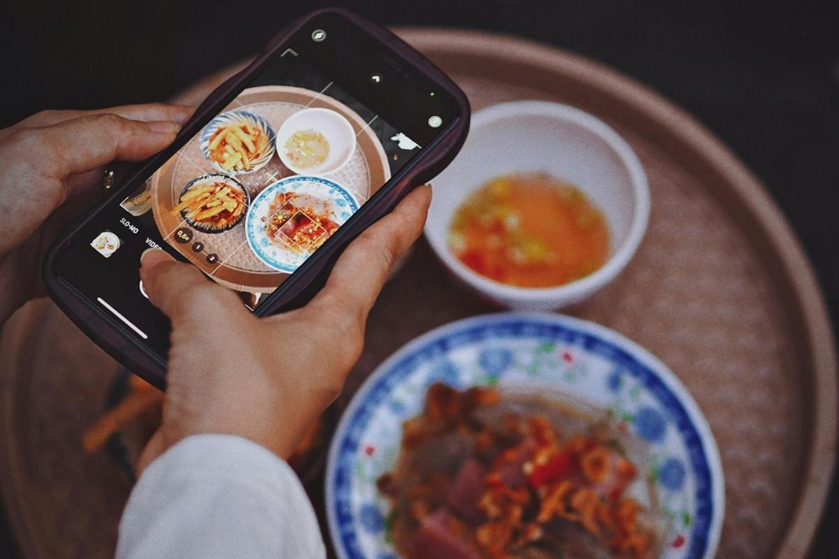 A top-down view shows a person holding a smartphone to take a photo of a round tray of Vietnamese food, including small plates of dishes and a bowl of dipping sauce. The phone screen displays a sharp, focused preview of the entire meal, while the actual food on the tray below remains softly blurred in the background. A top-down view shows a person holding a smartphone to take a photo of a round tray of Vietnamese food, including small plates of dishes and a bowl of dipping sauce. The phone screen displays a sharp, focused preview of the entire meal, while the actual food on the tray below remains softly blurred in the background.