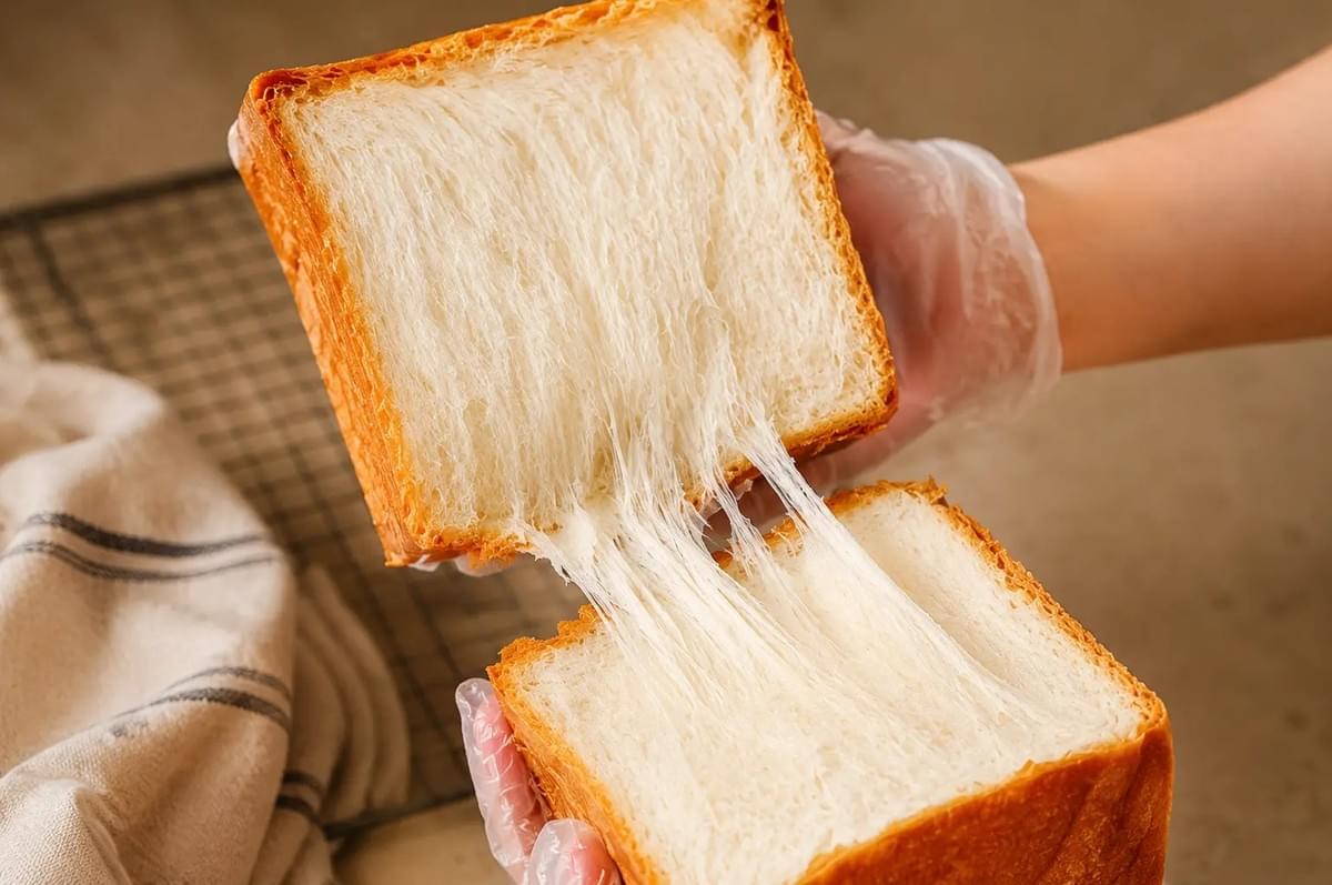 Gloved hands pulling apart a square slice of bread, showing fine, stretchy crumb strands. Gloved hands pulling apart a square slice of bread, showing fine, stretchy crumb strands.