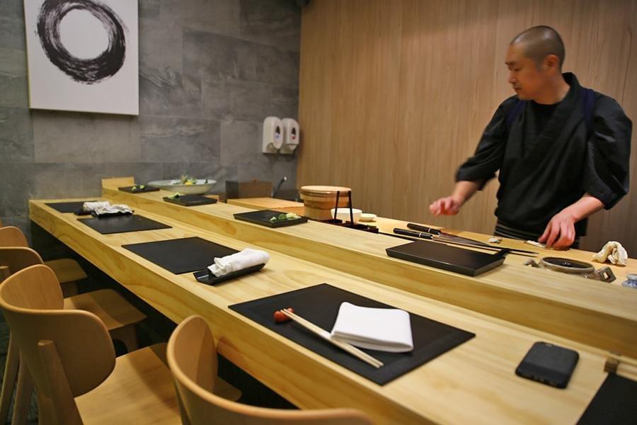 A Japanese sushi chef in dark attire works behind a minimalist light-wood counter set with place settings and wooden chairs. The sleek interior features grey tiled walls adorned with a framed black-and-white calligraphic circle artwork. A Japanese sushi chef in dark attire works behind a minimalist light-wood counter set with place settings and wooden chairs. The sleek interior features grey tiled walls adorned with a framed black-and-white calligraphic circle artwork.